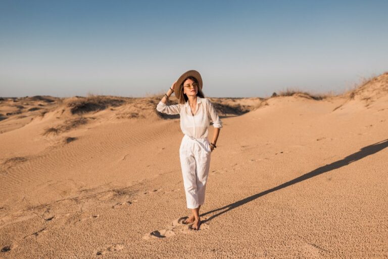 stylish-beautiful-woman-walking-desert-sand-white-outfit-wearing-straw-hat-sunset